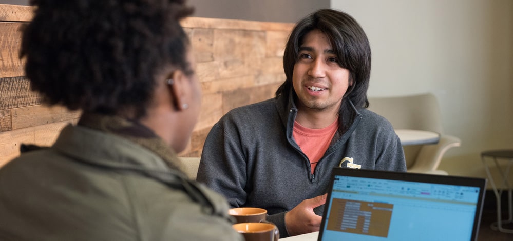 Two students talking while one looks at a spreadsheet on a computer